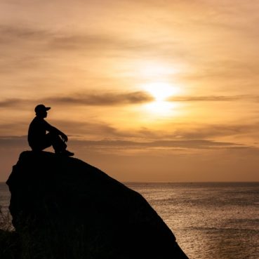 Silhouette man sitting on big rock with sightseeing of sunset in tropical sea